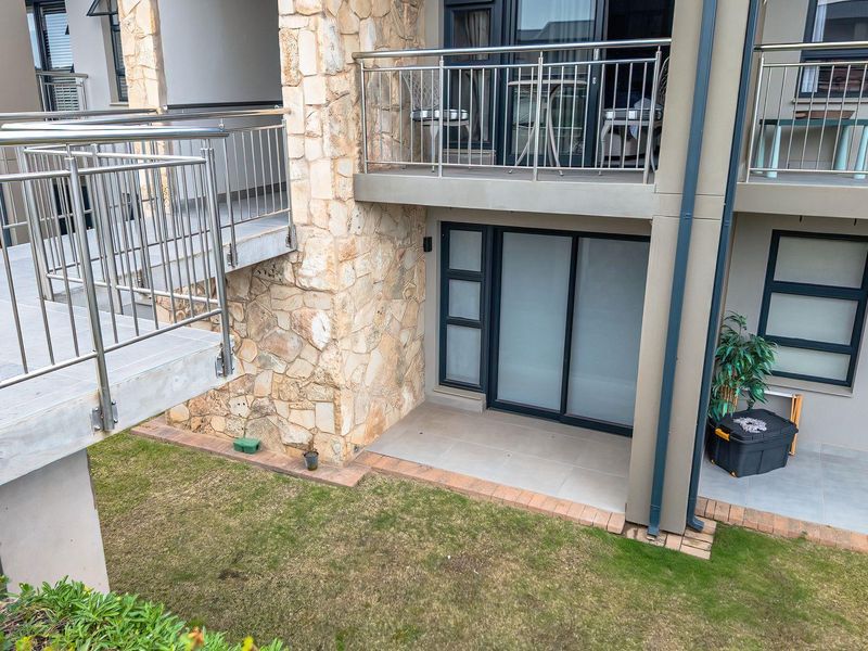 Rear view of a retirement village unit featuring a ground-level tiled patio, stone cladding accents, and upper-level balconies with stainless steel railings.