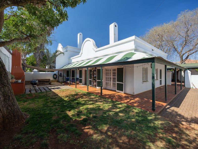 Rear view of the property featuring a covered patio with tiled flooring, a built-in braai, and classic gabled architecture set within a garden.