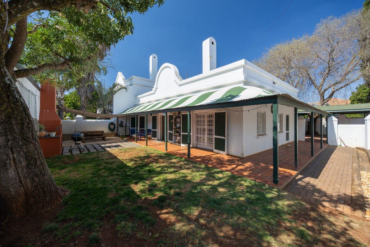 Rear view of the property featuring a covered patio with tiled flooring, a built-in braai, and classic gabled architecture set within a garden.