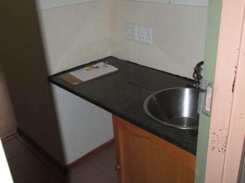A compact scullery or kitchenette area featuring built-in wooden cabinetry, a dark laminate countertop, a stainless steel sink, and tiled splashback.