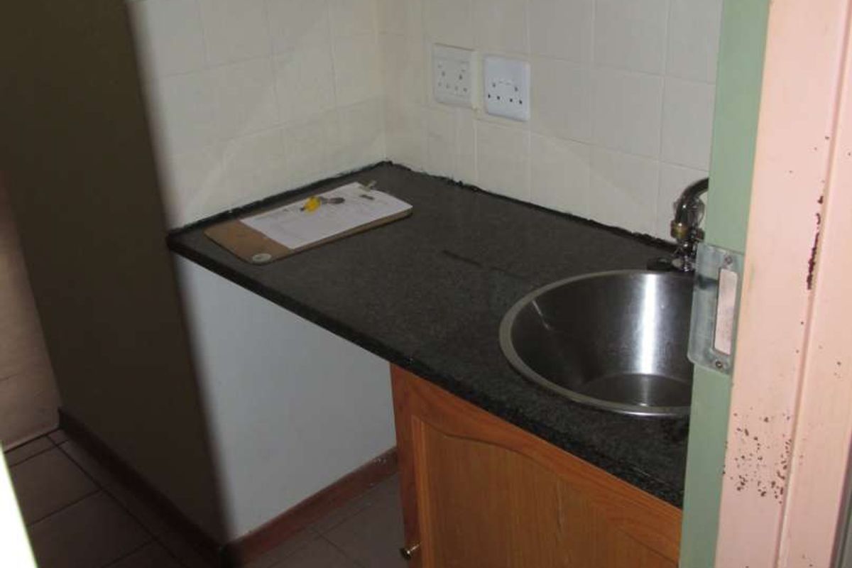 A compact scullery or kitchenette area featuring built-in wooden cabinetry, a dark laminate countertop, a stainless steel sink, and tiled splashback.
