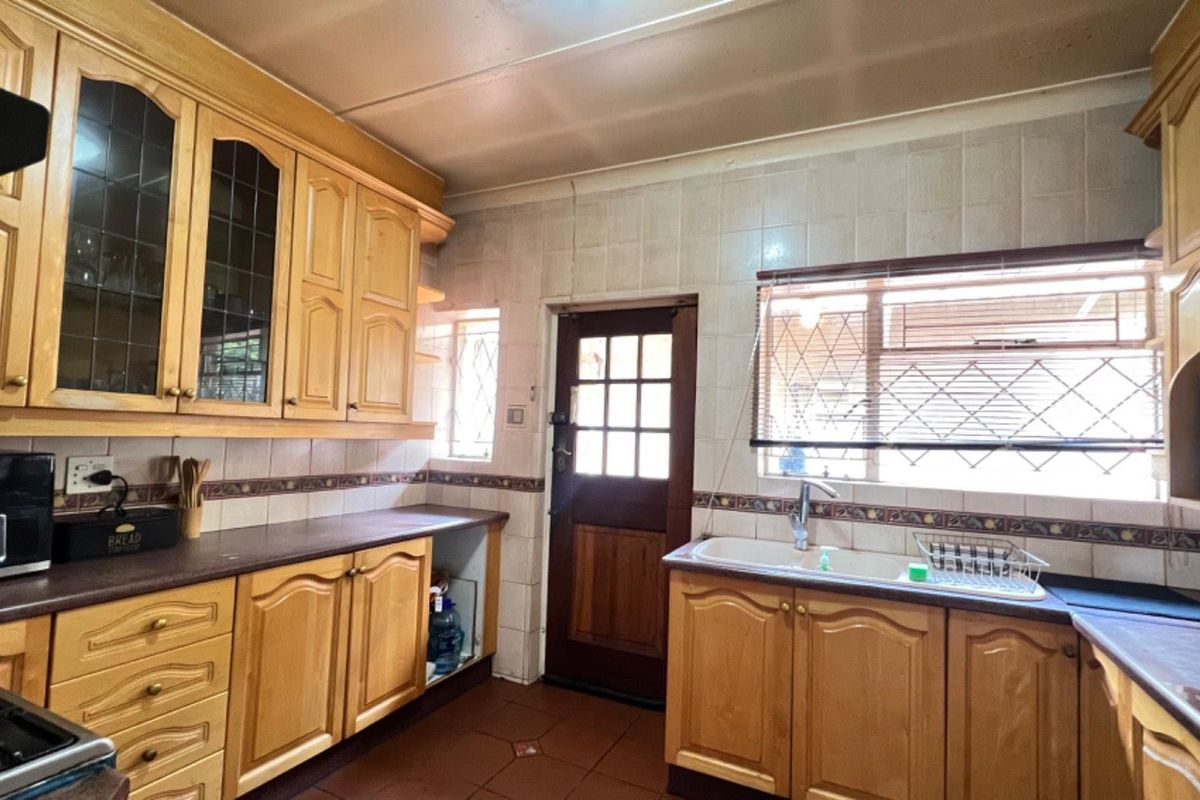 A functional kitchen featuring traditional timber cabinetry, tiled countertops, a double basin sink, and direct access to the exterior via a wooden door.