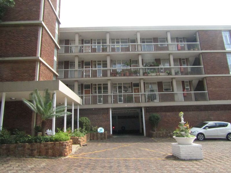 Front elevation of a multi-story brick apartment building featuring private balconies, a paved driveway, and covered ground-level parking access.