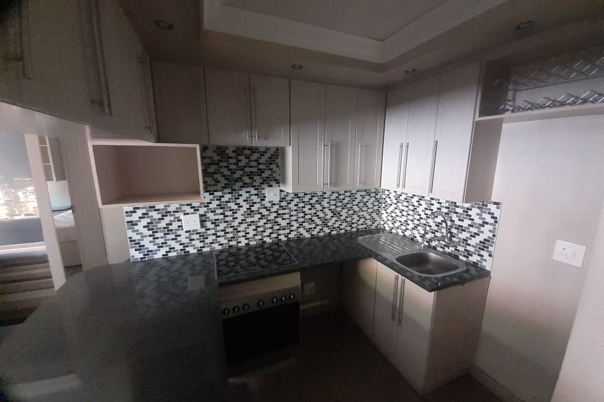 A compact, modern kitchen featuring light-toned cabinetry, a mosaic tile backsplash, stone countertops, and integrated appliances including an oven and hob.