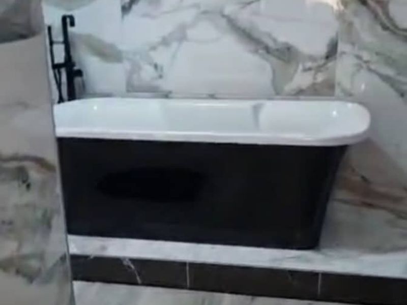 A modern bathroom featuring a sleek black freestanding bathtub set against elegant marble-look wall tiles and wood-effect flooring.