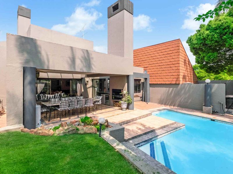 Outdoor entertainment area featuring a covered patio with a built-in braai, overlooking a swimming pool and landscaped garden.
