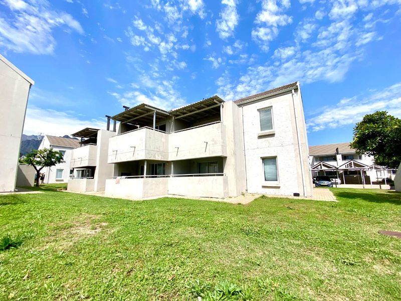 Rear view of a multi-story apartment complex featuring private balconies, paved pathways, and a shared lawn area under a clear blue sky.