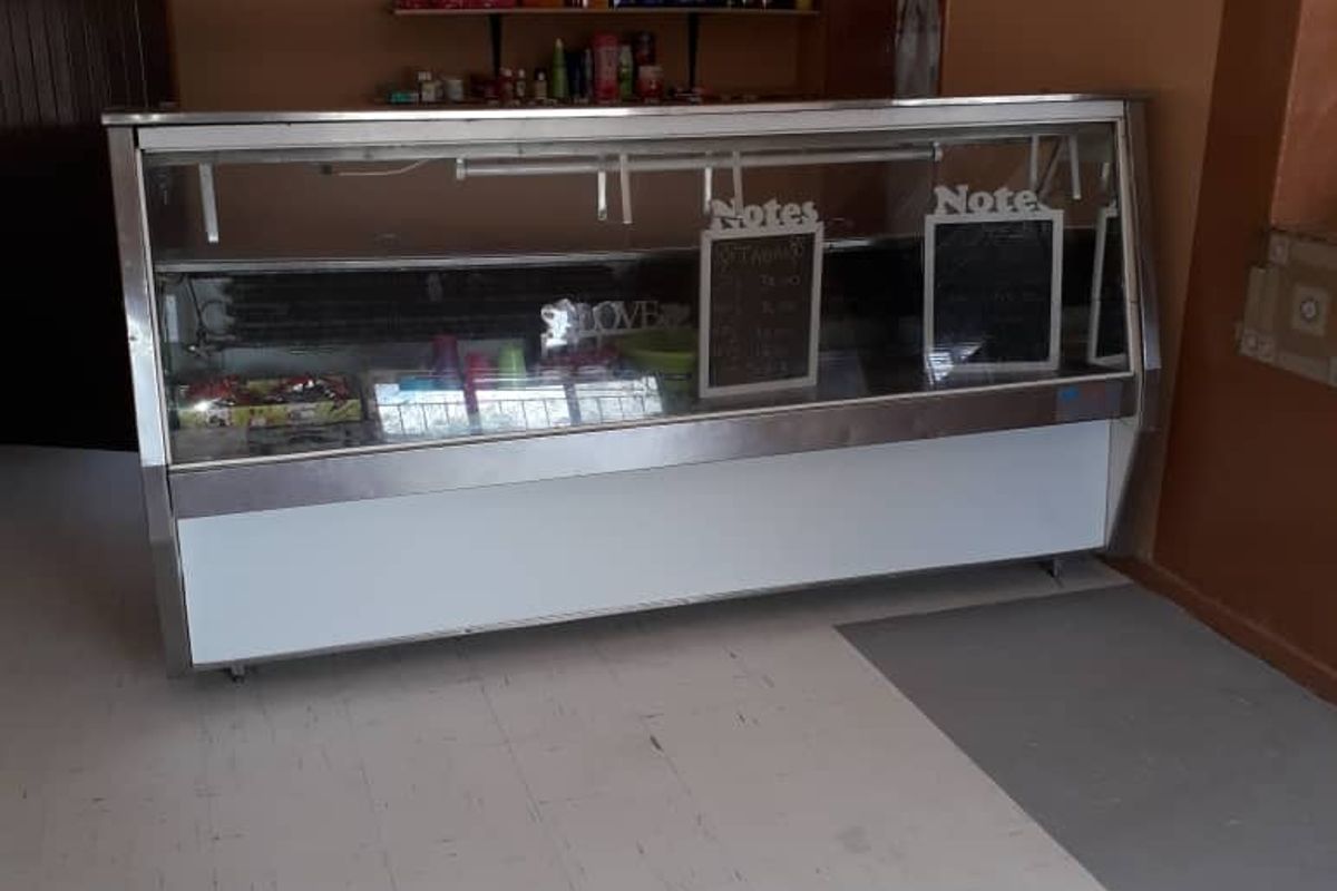 A commercial-style kitchen area featuring a large display fridge, wall-mounted shelving, and vinyl flooring, suitable for retail or food service use.