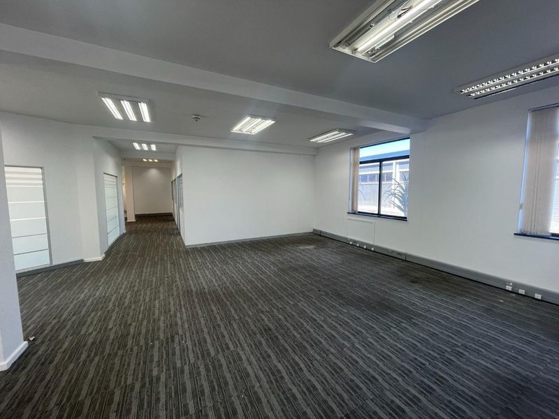 Spacious, empty open-plan office area featuring neutral walls, carpeted flooring, and ample natural light through large windows.