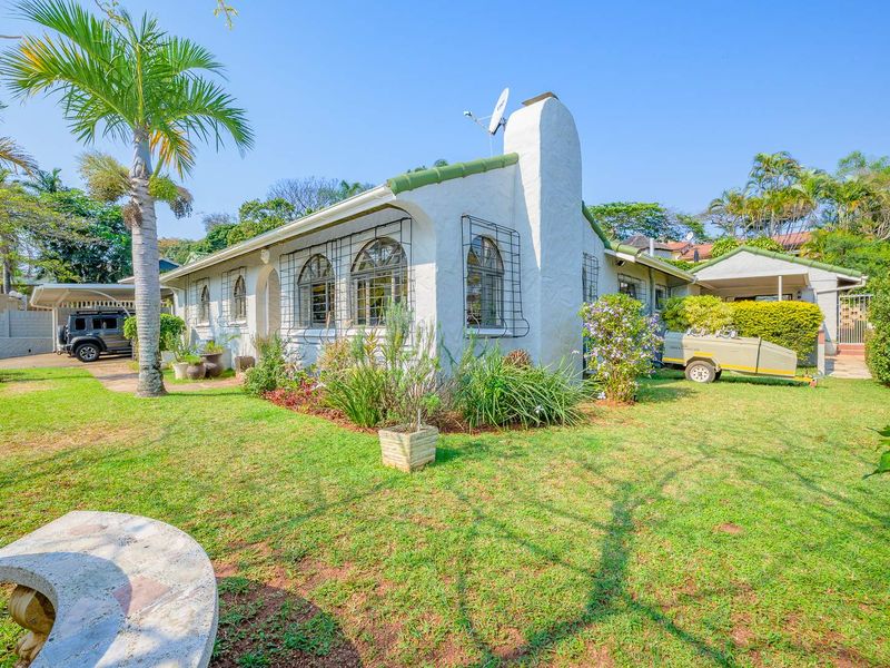 Rear view of a charming cottage-style house featuring arched windows, a prominent chimney, and a spacious garden with a paved driveway and carport.