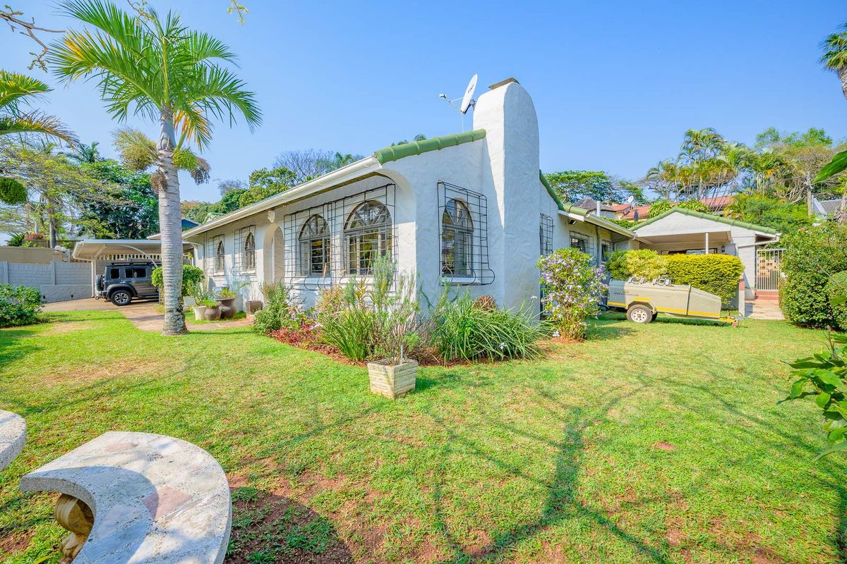 Rear view of a charming cottage-style house featuring arched windows, a prominent chimney, and a spacious garden with a paved driveway and carport.