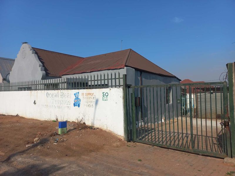 Front exterior view of a property featuring a metal gate, security fencing, and a corrugated metal roof with a prominent parapet wall.