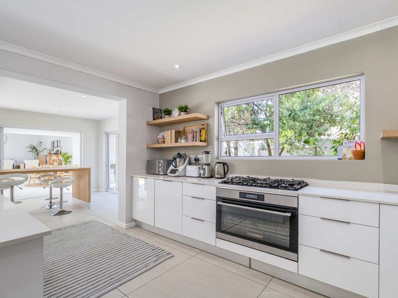 Modern kitchen featuring sleek white cabinetry, stone countertops, built-in oven, gas hob, and floating timber shelving, seamlessly flowing into the adjacent dining area.