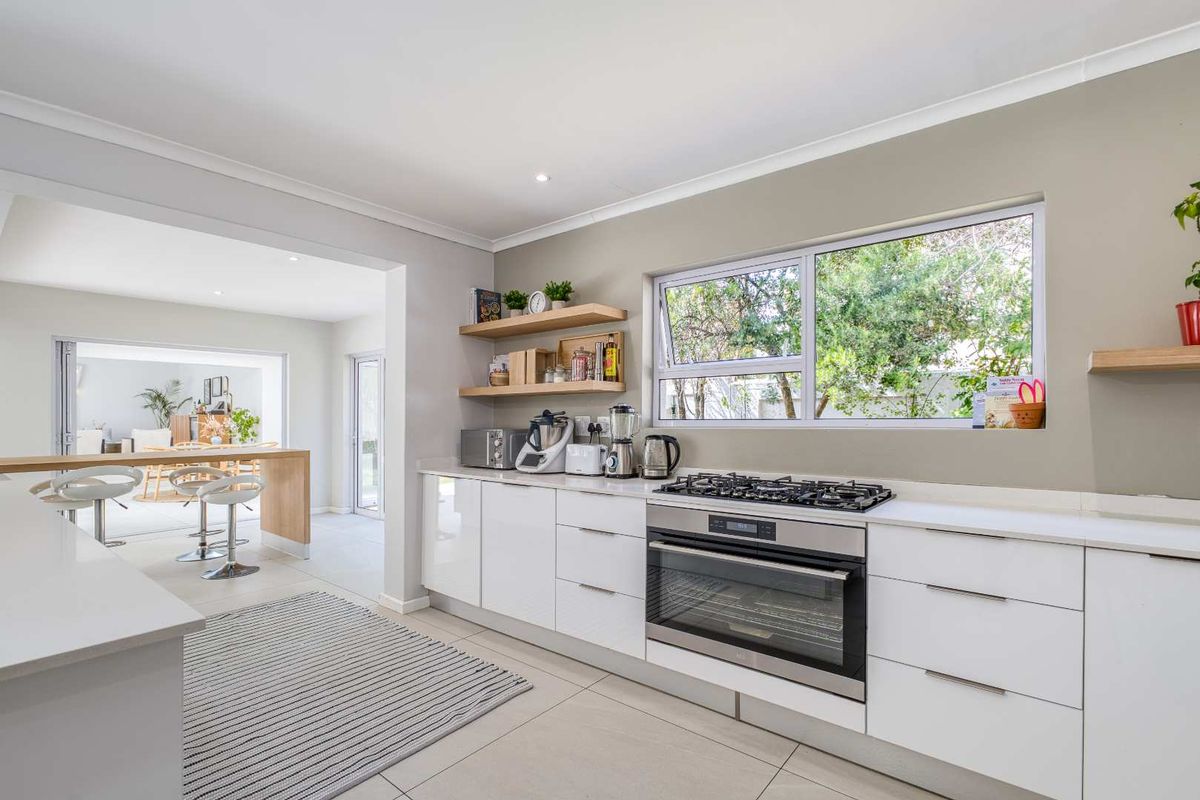 Modern kitchen featuring sleek white cabinetry, stone countertops, built-in oven, gas hob, and floating timber shelving, seamlessly flowing into the adjacent dining area.