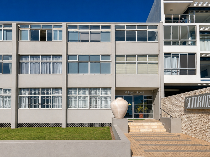 Front facade of the Sandringham apartment complex, featuring a clean concrete exterior, stone-clad signage wall, and paved entrance walkway.