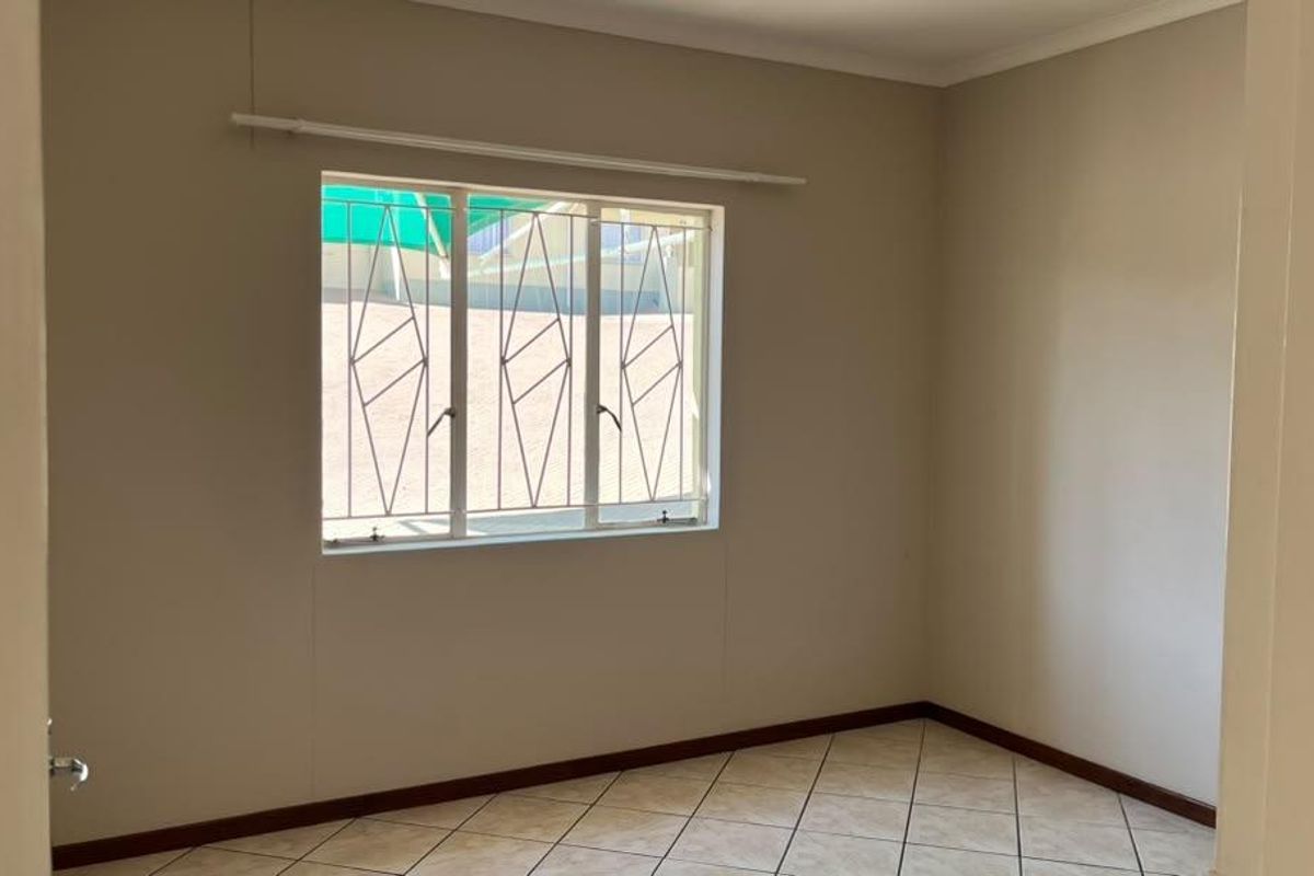 A clean, empty bedroom featuring neutral walls, tiled flooring, and a window fitted with security bars.