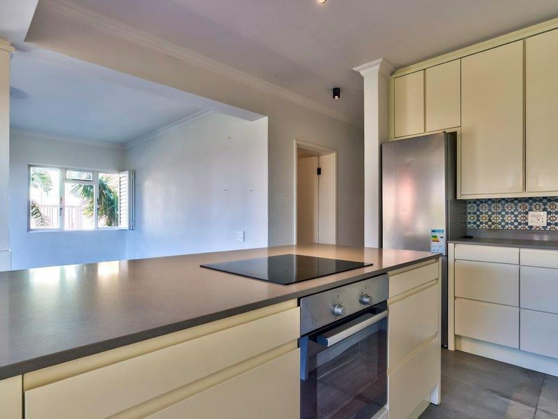 A clean, modern kitchen featuring built-in cabinetry, stone countertops, an integrated electric hob, and an oven, with an open-plan layout leading to the living area.
