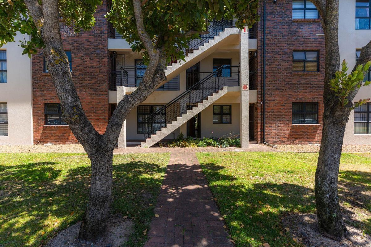 Rear view of a brick apartment building featuring an external staircase, metal railings, and a paved walkway leading through a landscaped garden area.