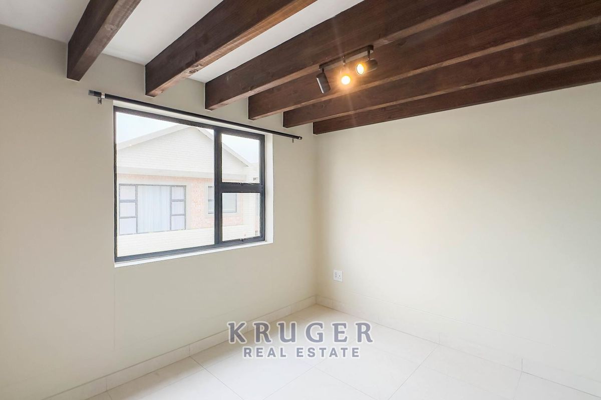 A bright, empty bedroom featuring exposed timber ceiling beams, modern track lighting, and neutral tiled flooring with an aluminium-framed window.