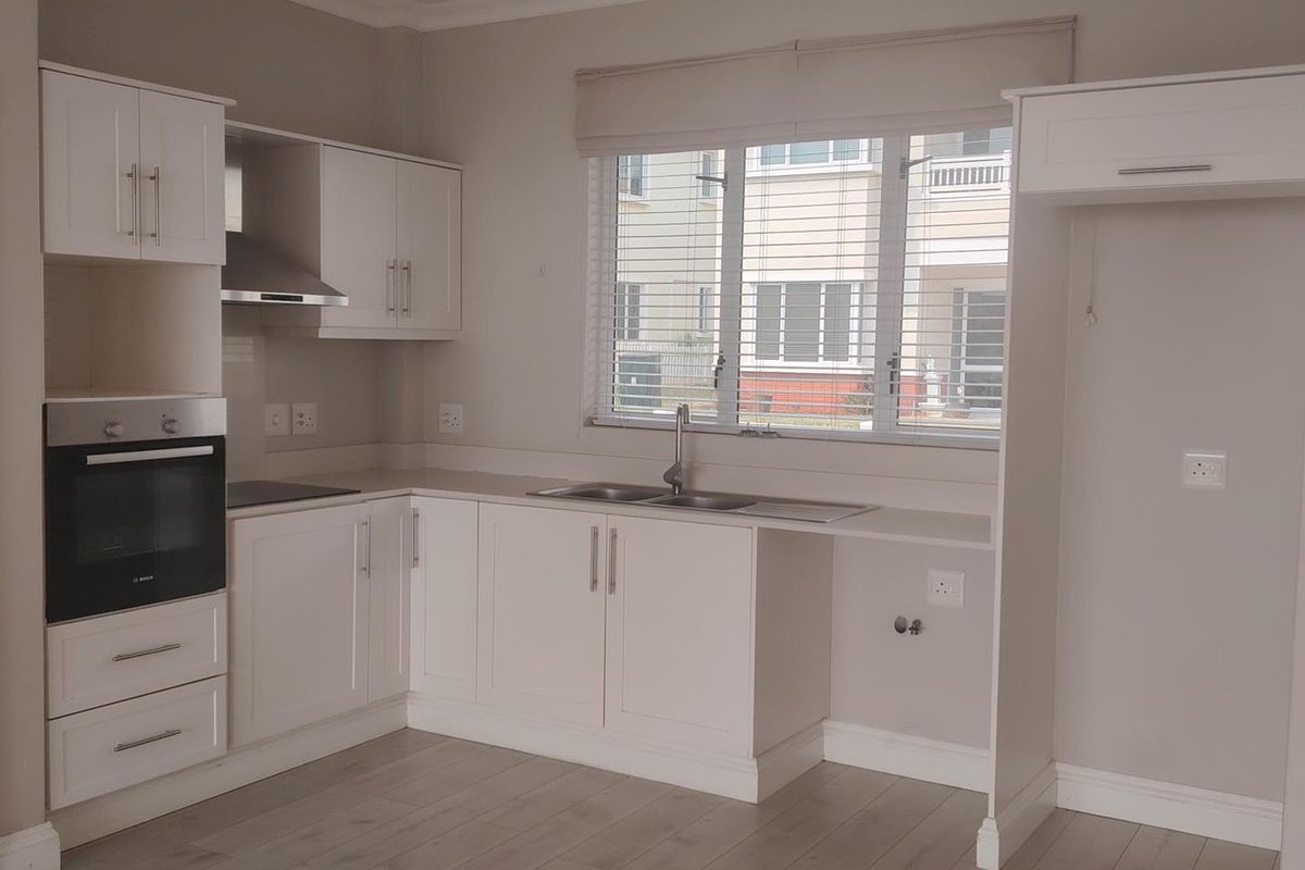 A clean, modern kitchen featuring white cabinetry, built-in oven, extractor fan, and stainless steel sink with neutral laminate flooring.