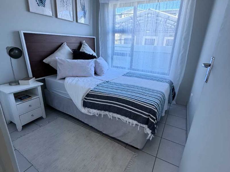 A compact, well-maintained bedroom featuring neutral tiled flooring, a large sliding window for natural light, and a built-in ventilation grille.