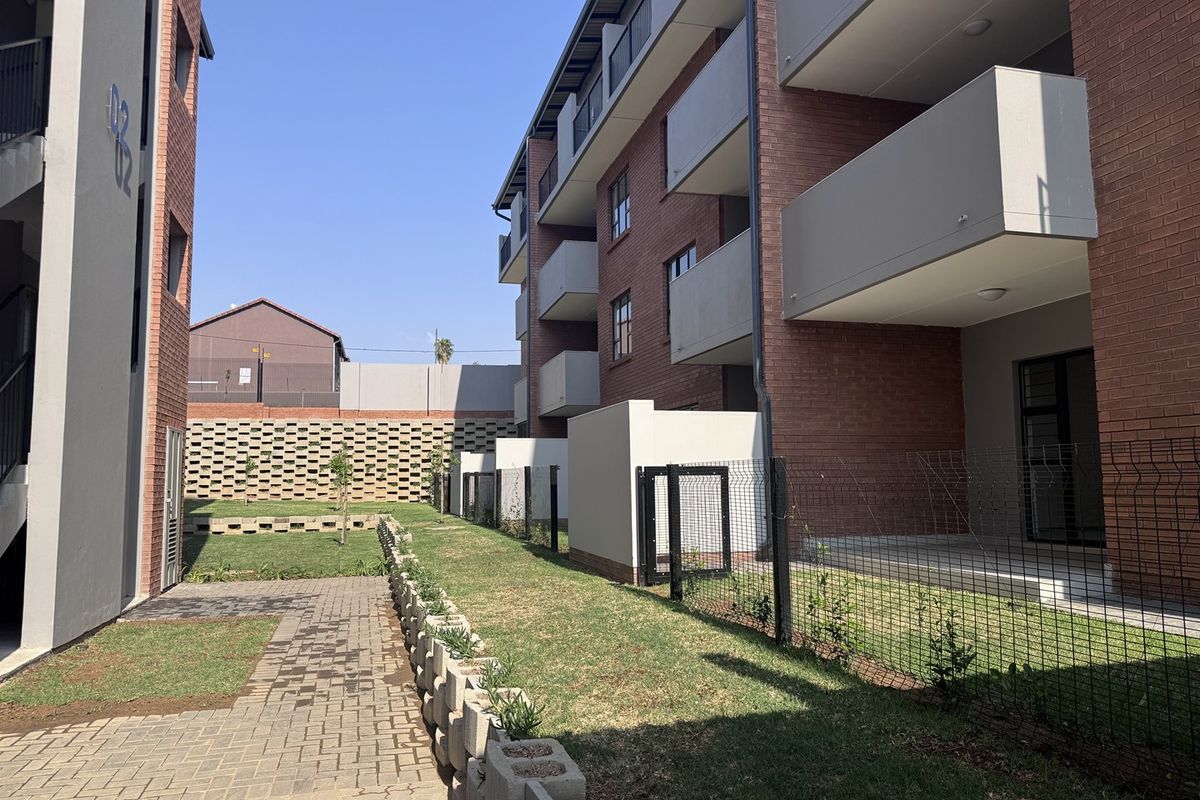 Exterior view of a modern apartment complex featuring brick facades, private balconies, a paved walkway, and a landscaped garden area with secure fencing.