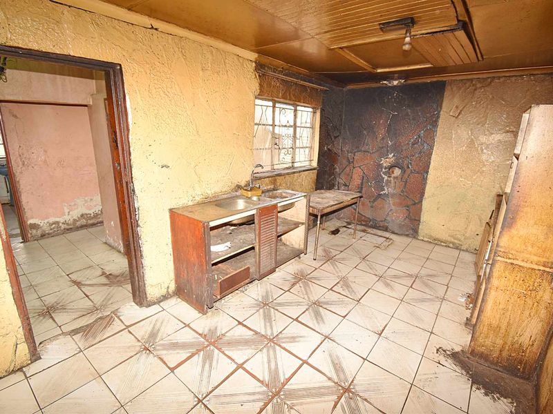 A dilapidated kitchen space requiring significant renovation, featuring original tiled flooring, basic sink unit, and extensive wall and ceiling damage.