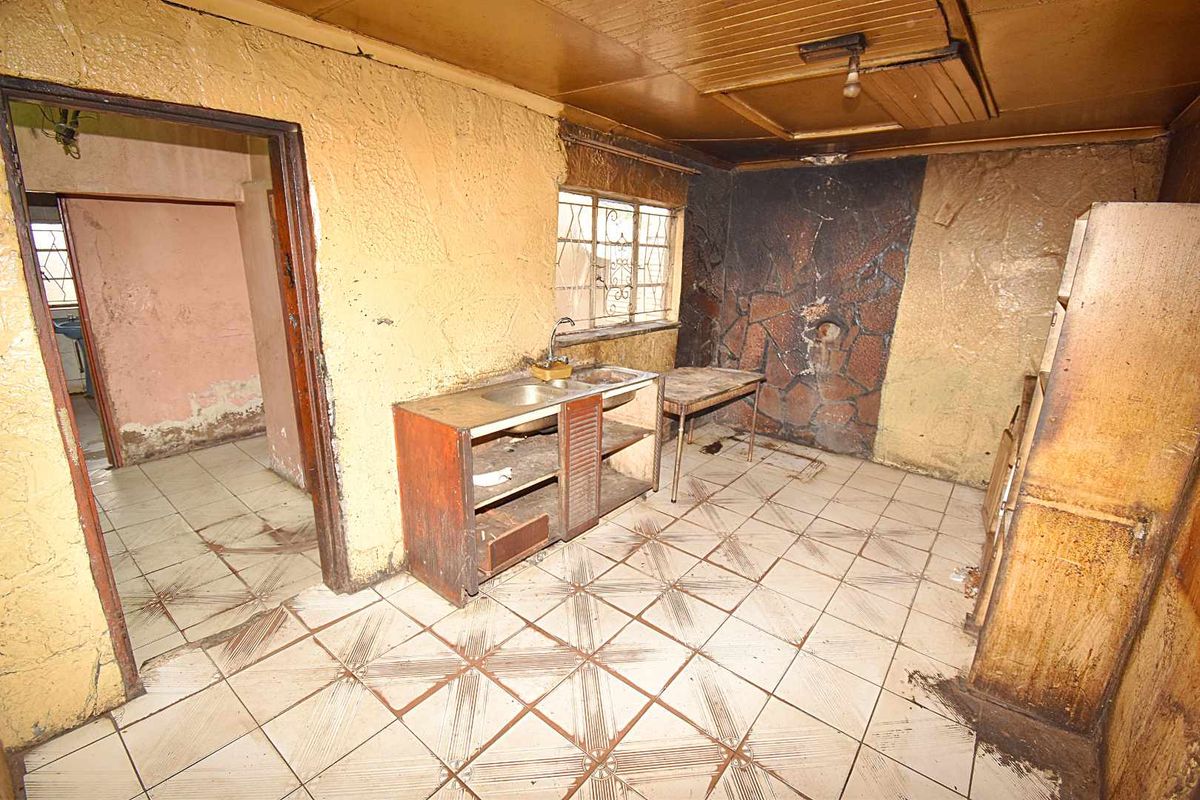 A dilapidated kitchen space requiring significant renovation, featuring original tiled flooring, basic sink unit, and extensive wall and ceiling damage.