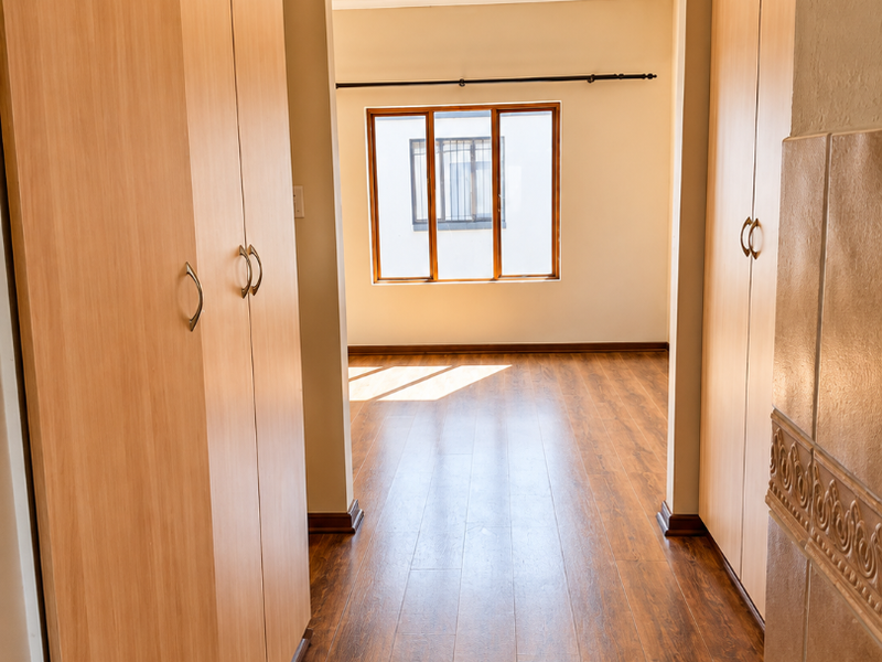 A bright, empty bedroom featuring light wood-finish built-in cupboards, warm-toned laminate flooring, and a window with security bars.