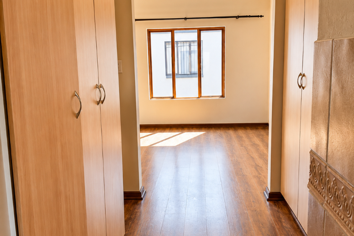 A bright, empty bedroom featuring light wood-finish built-in cupboards, warm-toned laminate flooring, and a window with security bars.