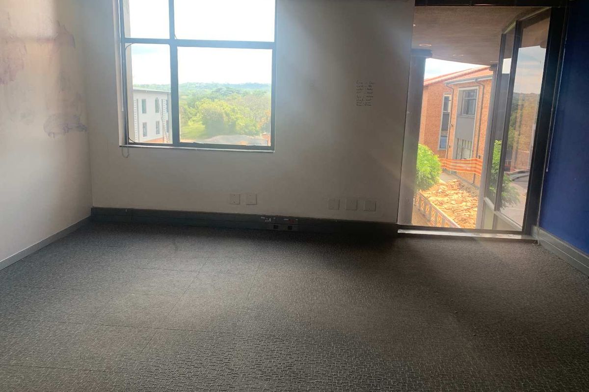 An empty office space featuring large windows, carpeted flooring, and a suspended ceiling, offering a bright workspace with views of the surrounding area.