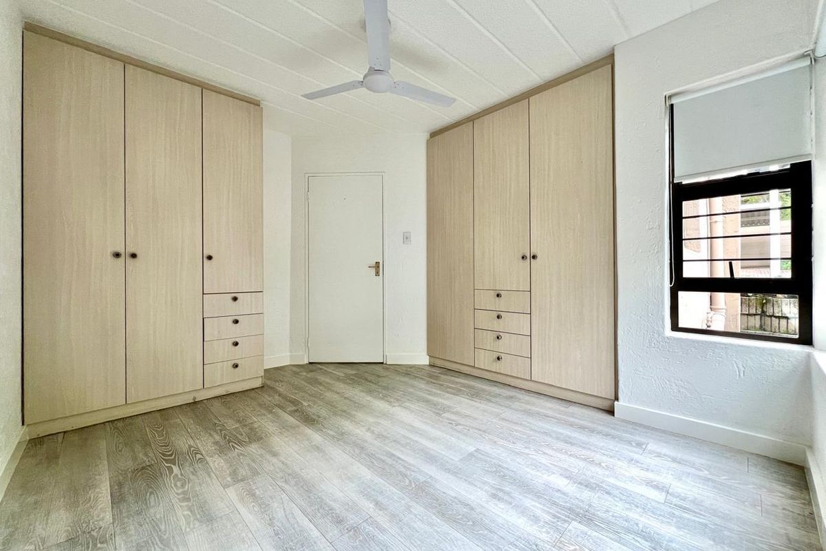 A bright bedroom featuring light wood-finish built-in cupboards, modern wood-look flooring, a ceiling fan, and a window with security bars.