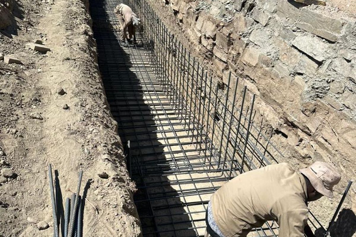 Construction site showing steel reinforcement rebar cage installation for a foundation or retaining wall footing on a residential plot.