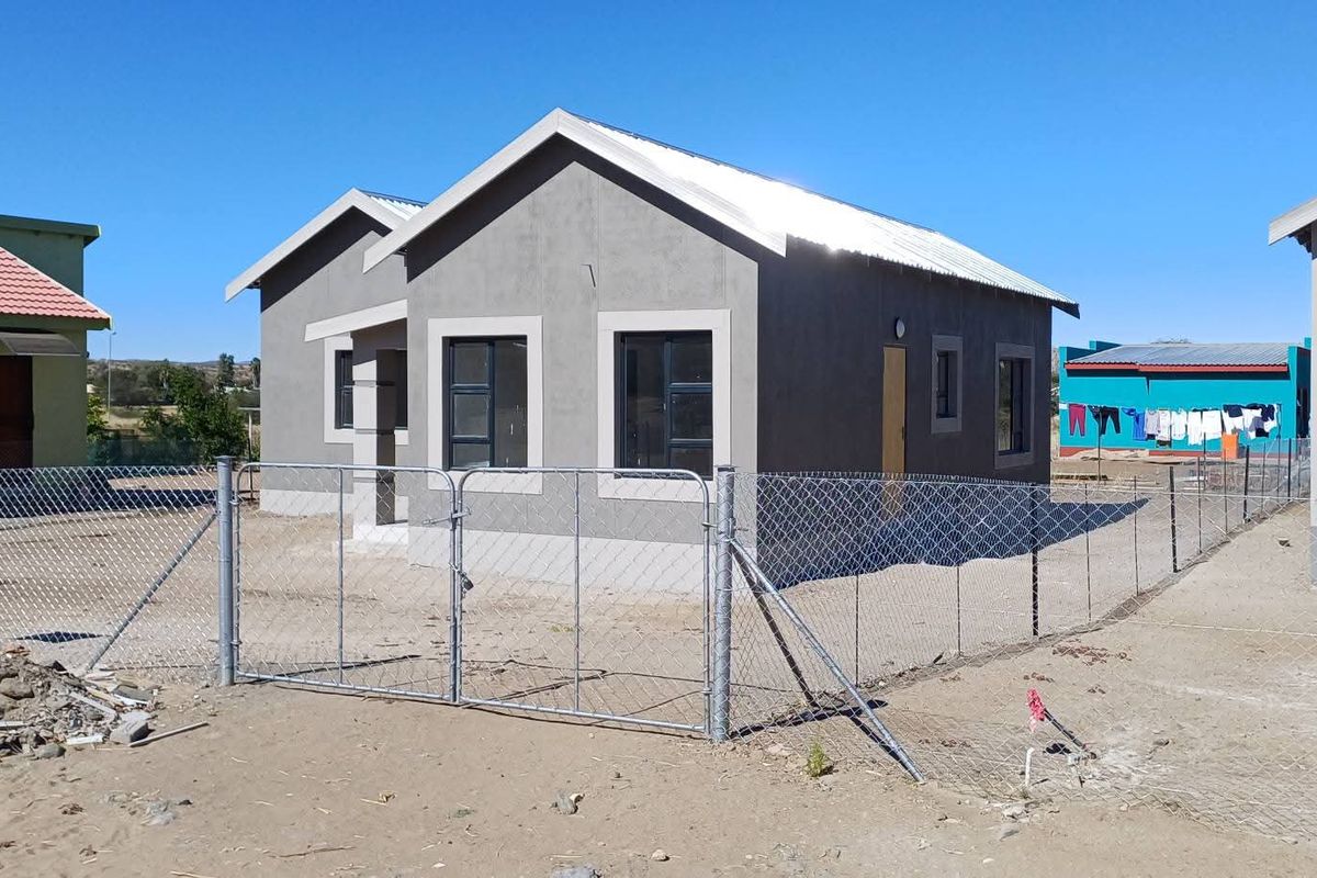 A newly constructed single-story house featuring a grey facade, metal roofing, and a perimeter chain link fence in a developing residential area.