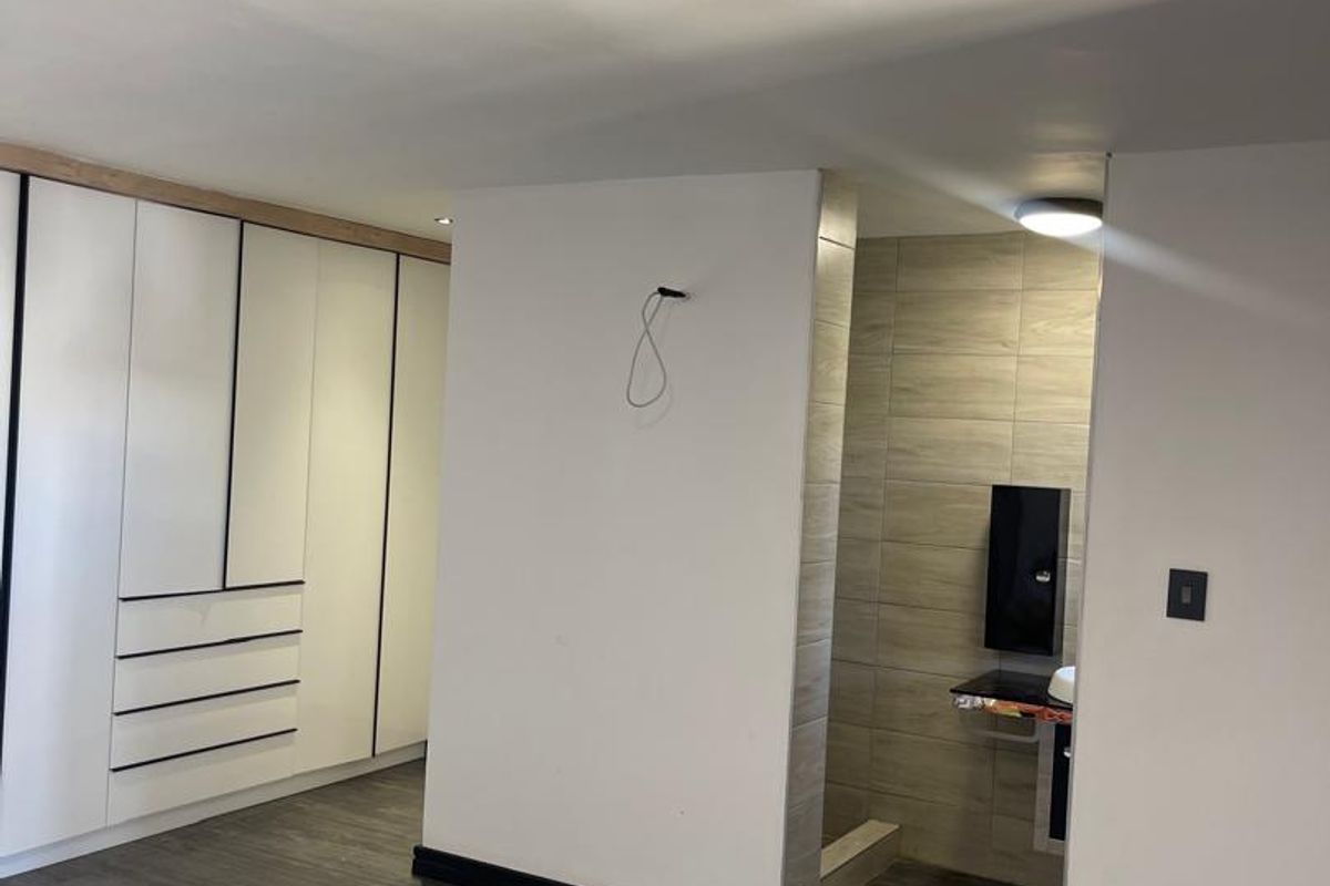 A modern bedroom featuring built-in cupboards, laminate flooring, and an open-plan ensuite area with tiled finishes.