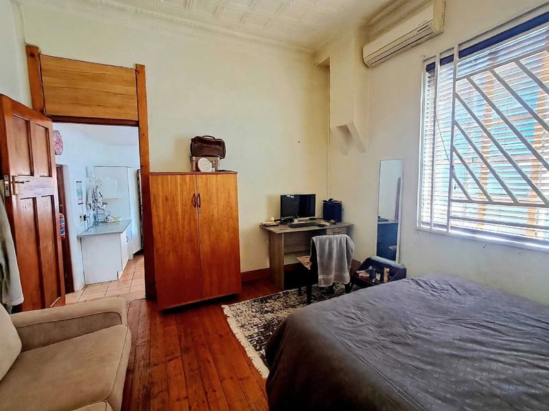 A bedroom featuring original pressed metal ceilings, hardwood flooring, and a wall-mounted air conditioning unit, with an adjoining view into a kitchen area.