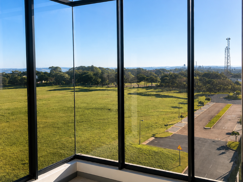 A bright, empty corner room featuring expansive floor-to-ceiling windows that offer panoramic views of the surrounding green landscape and parking area.