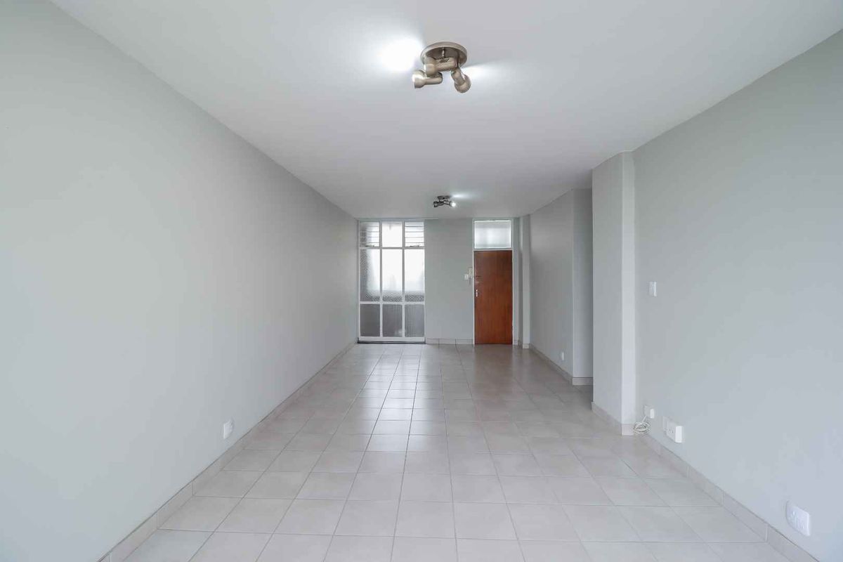 A spacious, empty living area featuring neutral tiled flooring, light-colored walls, and a functional entrance door with natural light from the adjacent window.