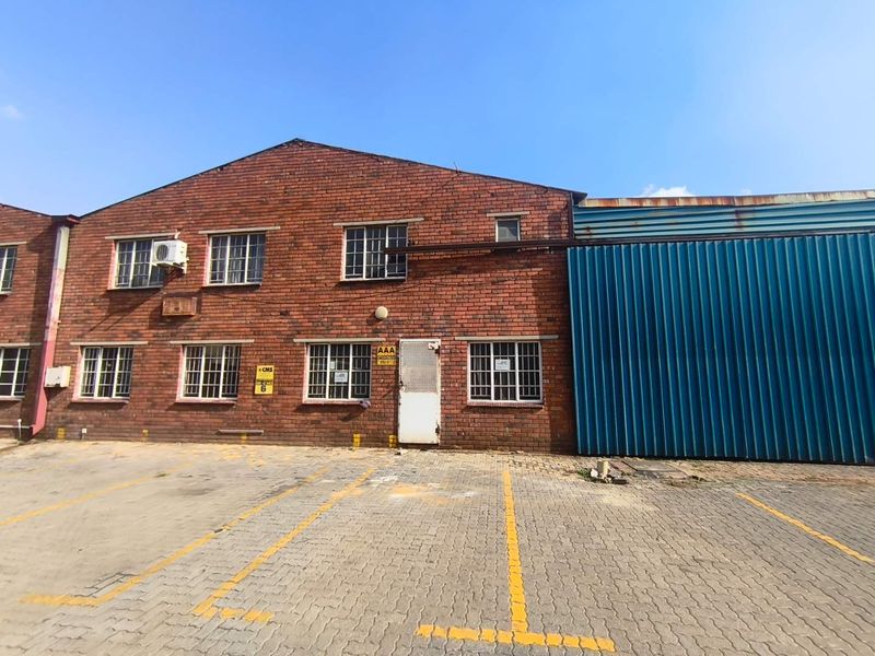 Industrial property exterior featuring a brick facade, corrugated metal siding, security-barred windows, and a paved parking area.