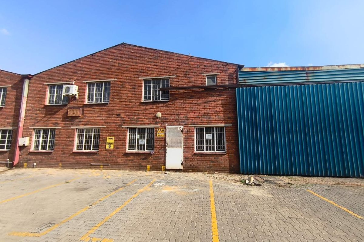 Industrial property exterior featuring a brick facade, corrugated metal siding, security-barred windows, and a paved parking area.
