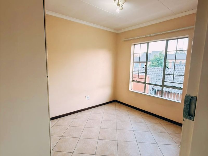 A clean, empty bedroom featuring neutral tiled flooring, a built-in cupboard, and a window with security bars, offering a bright and versatile living space.