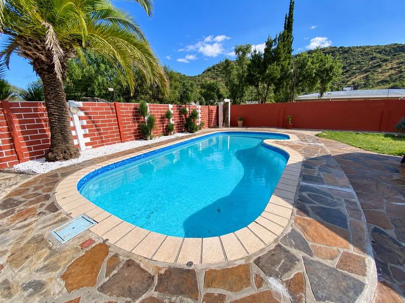 A well-maintained outdoor swimming pool area featuring stone paving, a brick boundary wall, and established landscaping with mountain views.