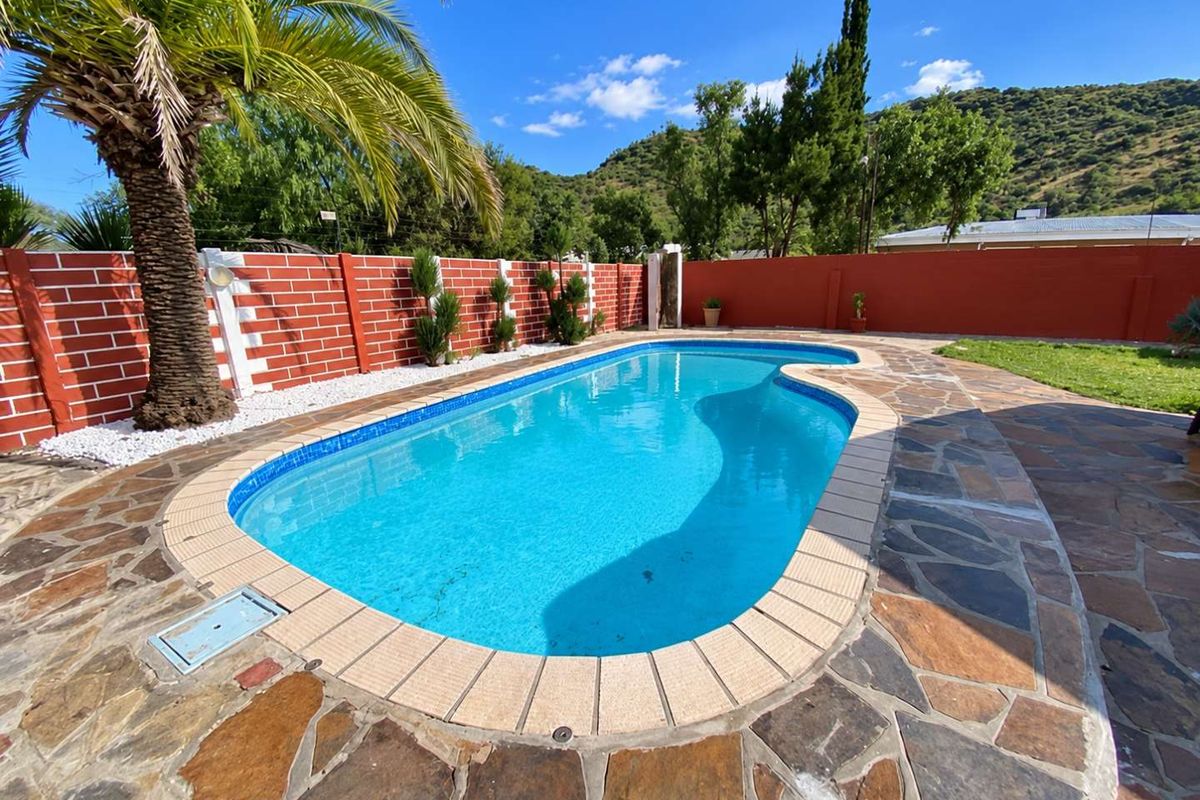 A well-maintained outdoor swimming pool area featuring stone paving, a brick boundary wall, and established landscaping with mountain views.