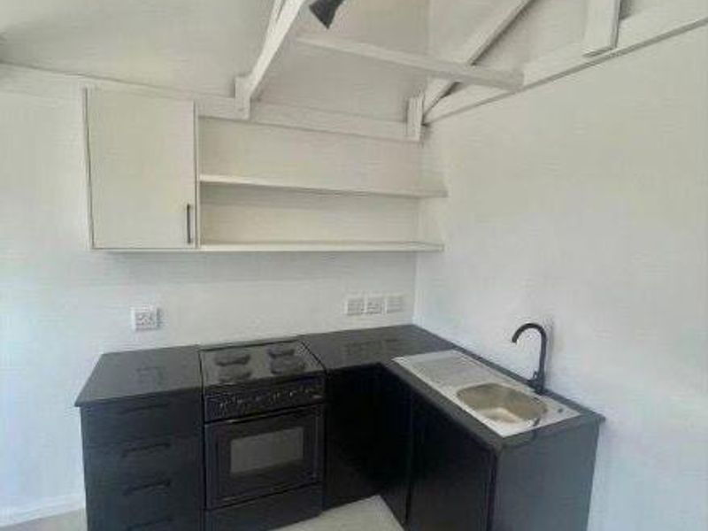 Compact kitchen featuring black cabinetry, built-in oven, electric hob, stainless steel sink, and open shelving under an exposed beam ceiling.