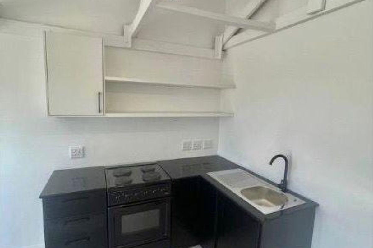 Compact kitchen featuring black cabinetry, built-in oven, electric hob, stainless steel sink, and open shelving under an exposed beam ceiling.