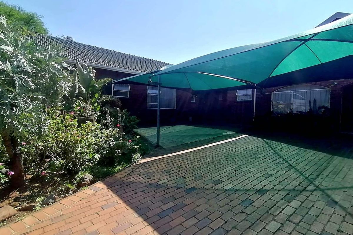 Rear exterior view featuring a brick-paved patio area covered by a green shade sail, adjacent to a landscaped garden and brick building facade.