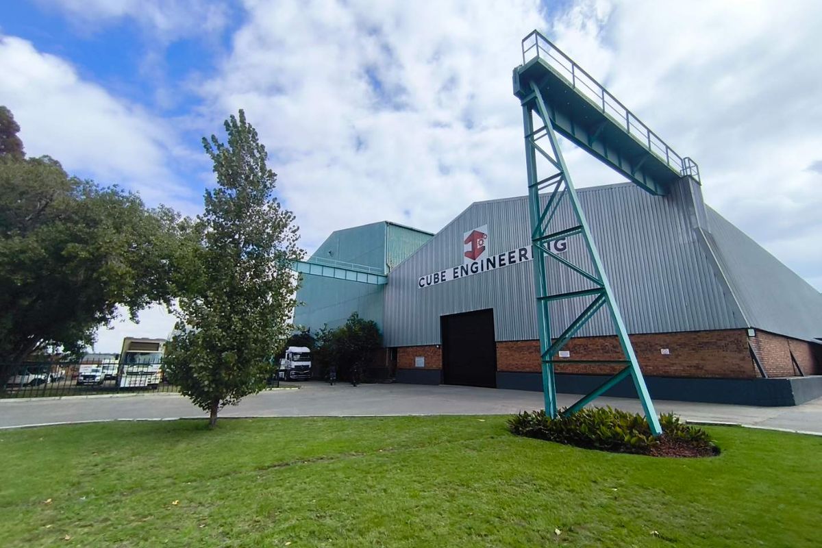 Front view of an industrial facility featuring metal cladding, brick base, and an external gantry crane structure set within a landscaped yard.