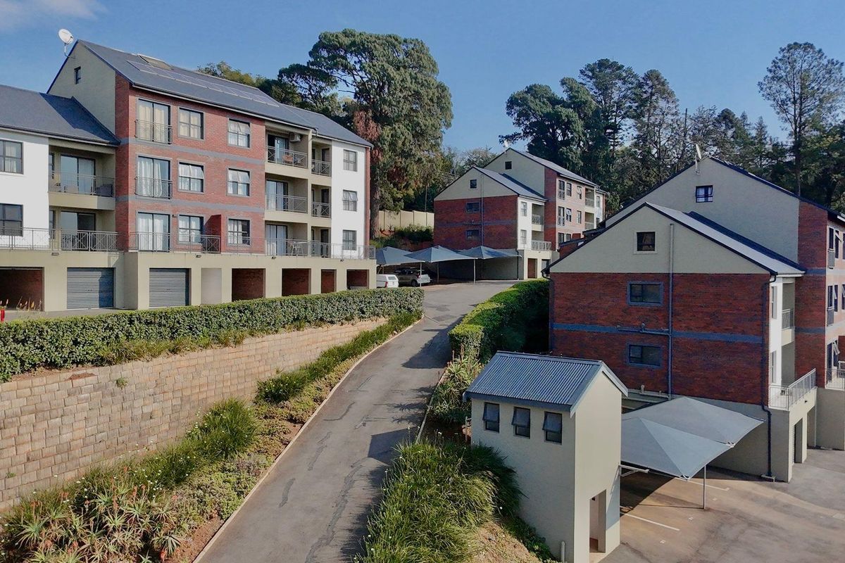 An elevated view of a multi-story apartment complex featuring brick and plaster facades, paved driveways, covered parking bays, and landscaped retaining walls.