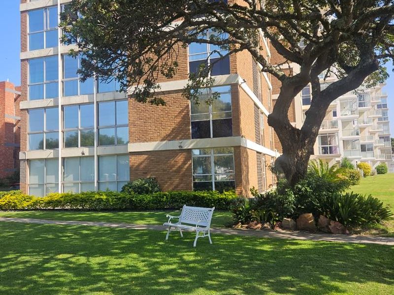 Exterior view of a multi-story apartment building featuring a brick facade, large windows, and a well-maintained garden area with a mature tree.