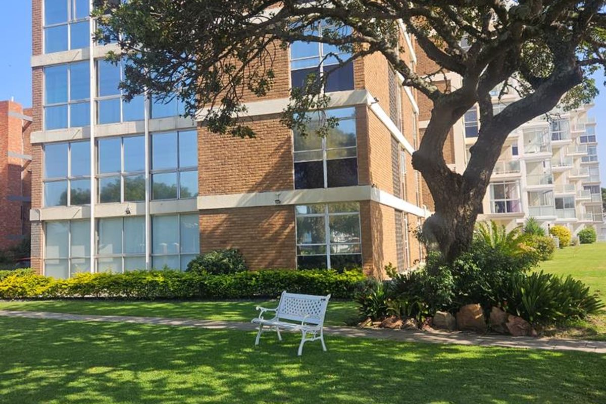 Exterior view of a multi-story apartment building featuring a brick facade, large windows, and a well-maintained garden area with a mature tree.